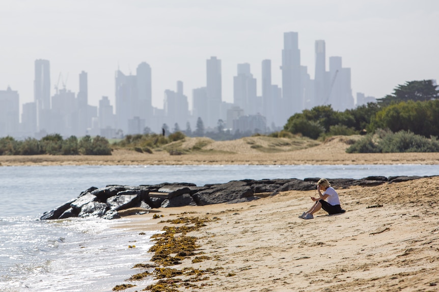 A woman sits on a beach with Melbourne city in the background.