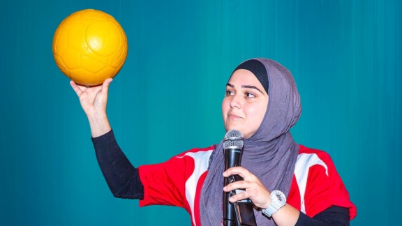 A woman wearing a hijab holds a microphone in one hand and a yellow football in another.