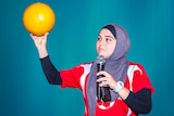 A woman wearing a hijab holds a microphone in one hand and a yellow football in another.