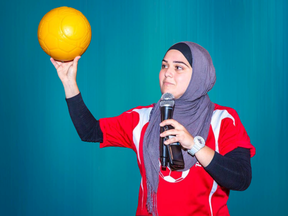 A woman wearing a headscarf holds up a soccer ball in one hand and a microphone in another.