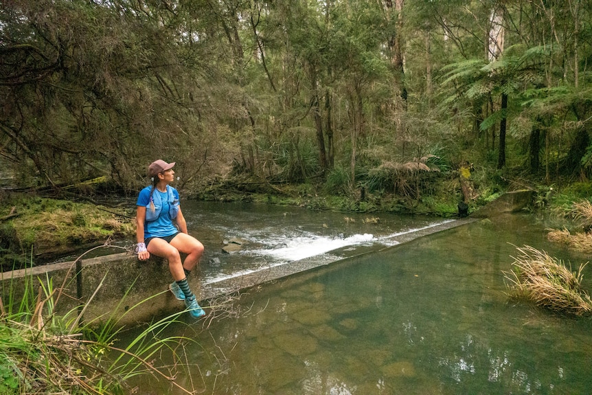 A woman sits next to a river surrounded by bushland.