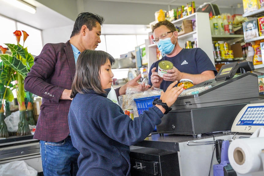 A young girl serves at a shop counter while a man standing next to her chats to a customer holding fruit.