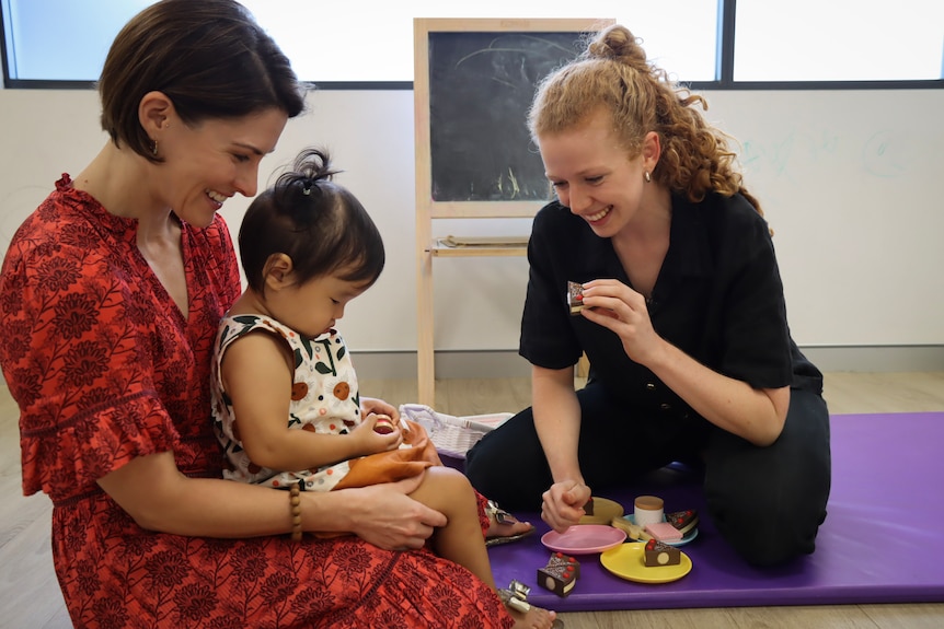 Two women and a toddler sit on a mat on the ground playing with toys and bubbles. Everyone is smiling.