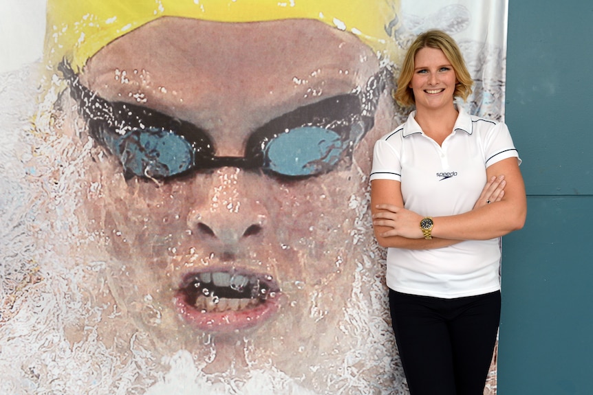 Female swimmer standing in front of a mural of her with her arms folded and smiling.