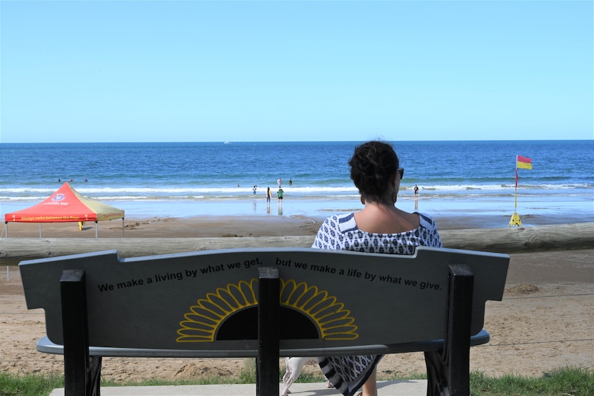 A lady in a dress sits on a bench looking at the beach with people playing in the shallow water.