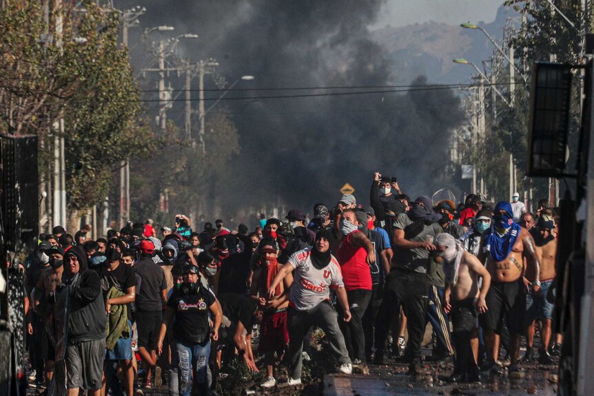 Protesters throw rocks at police as a plume of smoke rises in the distance