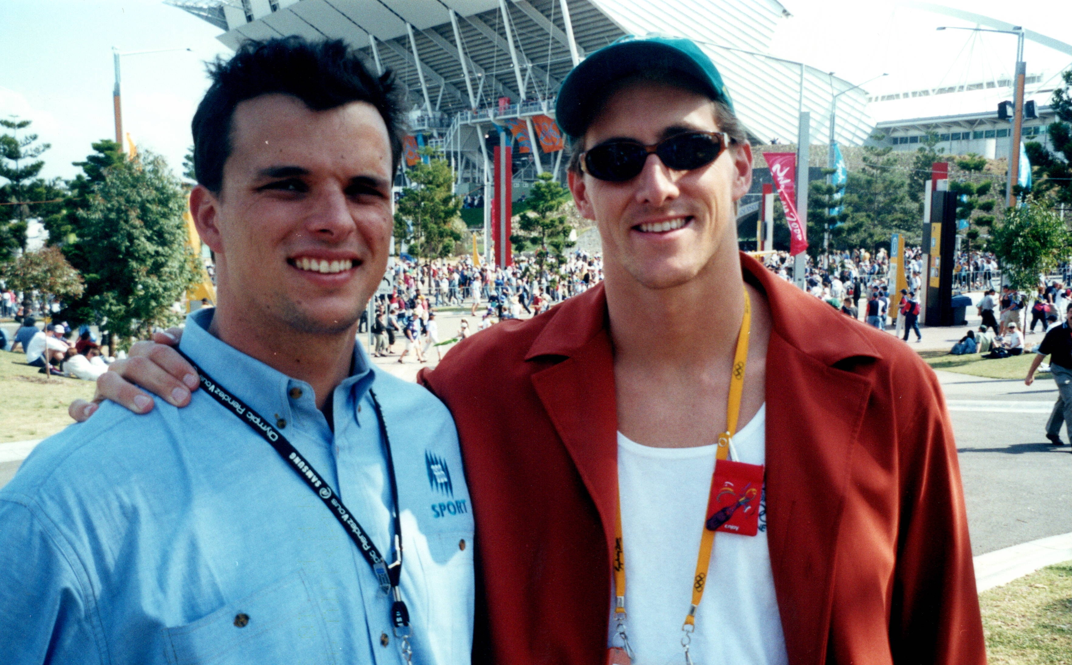 Two young men stand next to each other. One has a blue SBS polo on and the other has a cap, sunglasses and red shirt.