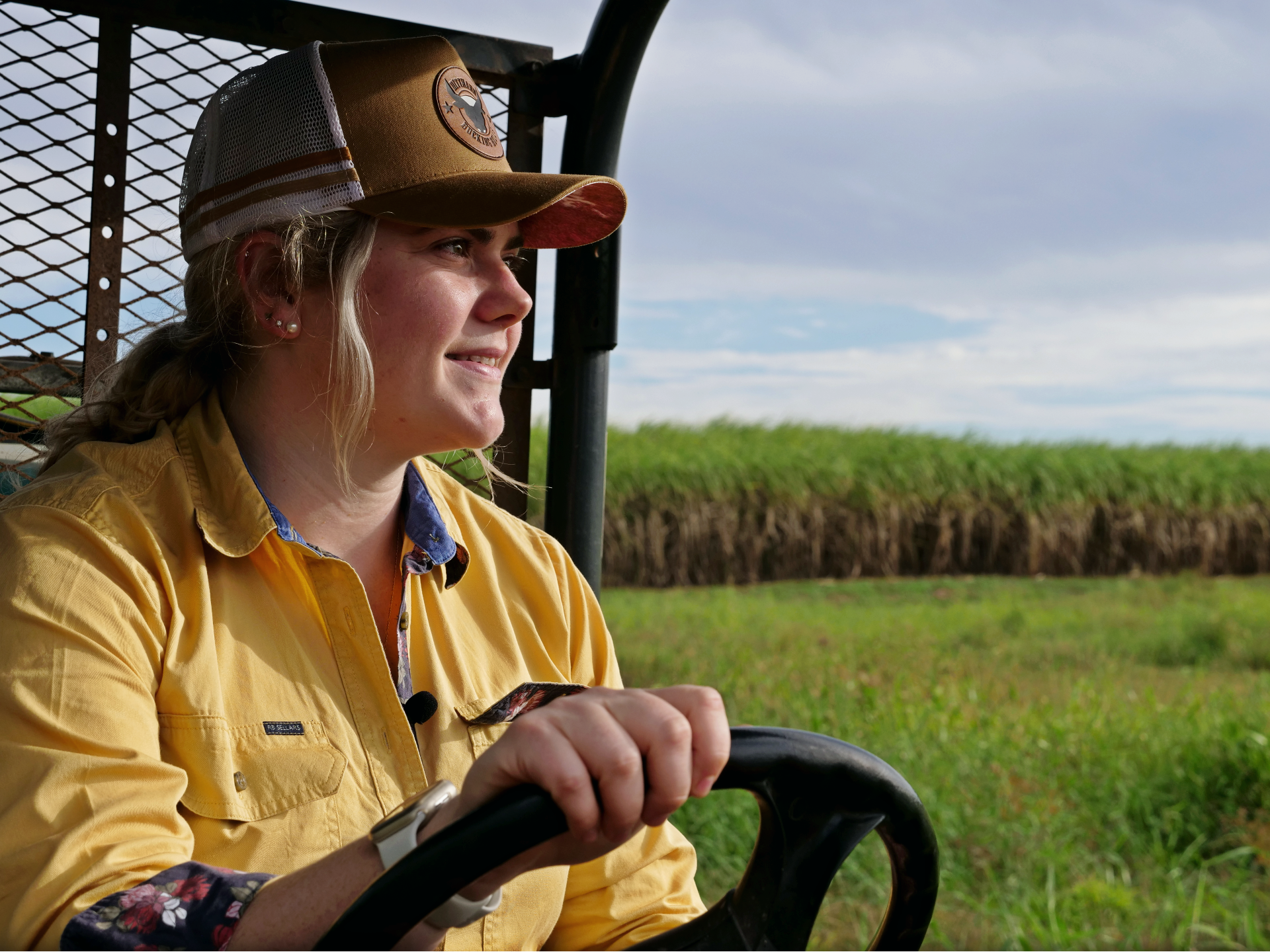 a young woman holds the steering wheel of a quad bike on a cane farm, smiling off into the distance