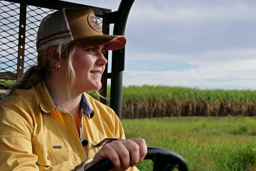 a young woman holds the steering wheel of a quad bike on a cane farm, smiling off into the distance