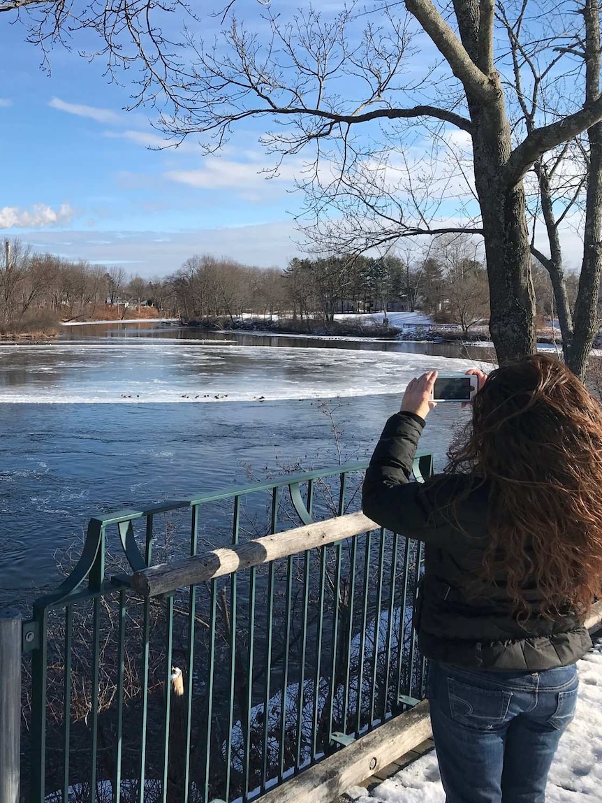 A woman stands on a river bank holding up her phone to capture a circular ice formation floating on the water