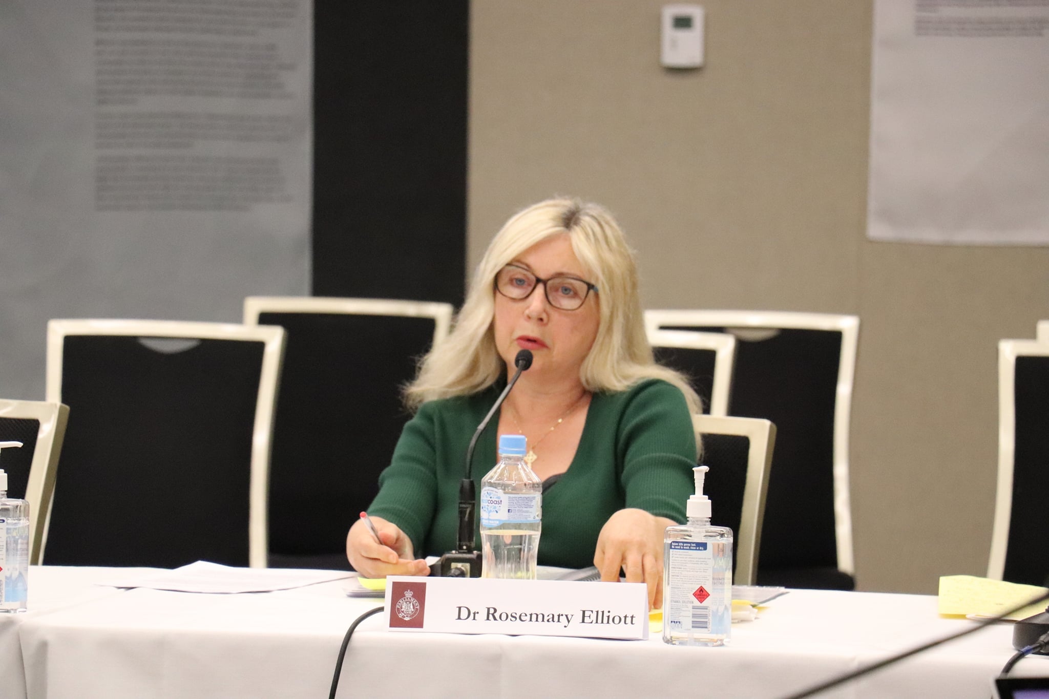A woman with long blonde hair and a dark green top sits at a desk behind a microphone.