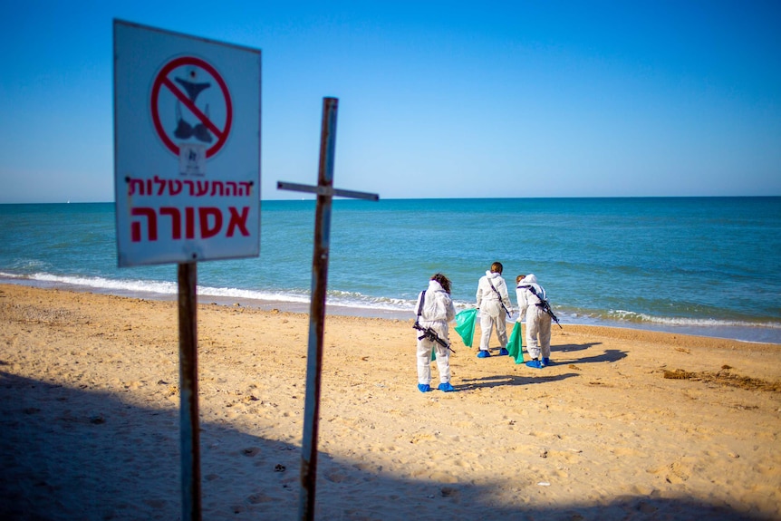 Three people in protective suits with rifles slung over their shoulders standing on a beach with plastic bags