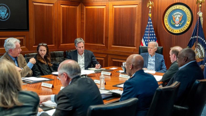 Joe Biden and others are seated around a large table in a wood-panelled room.
