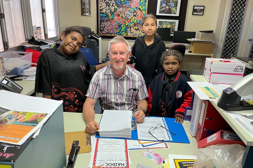 A man with grey-white hair sits at a desk and smiles at the camera, surrounded by three smiling young girls.