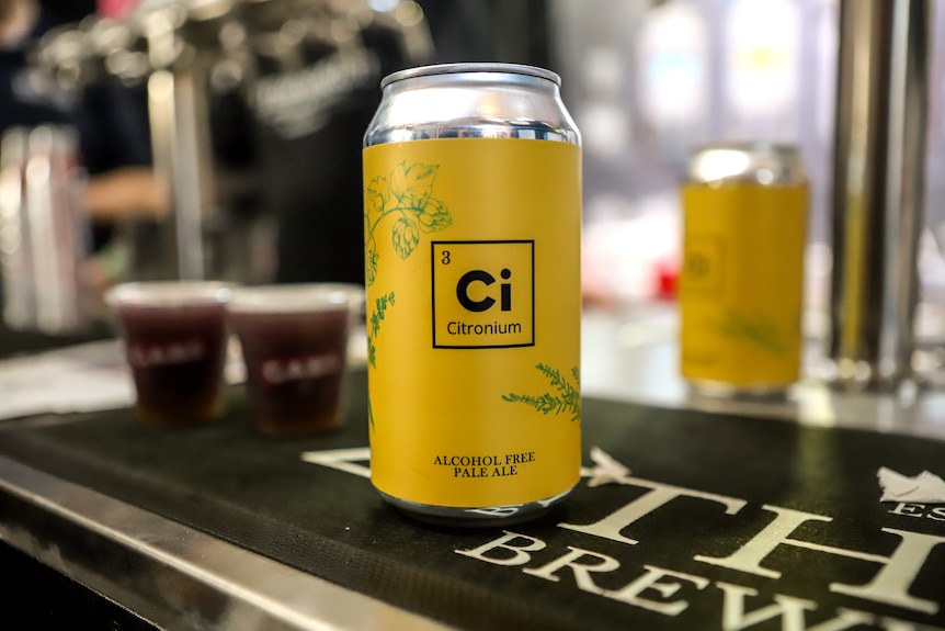 Yellow beer can sitting on a bar mat benchtop with a second can and two small plastic shot cups behind