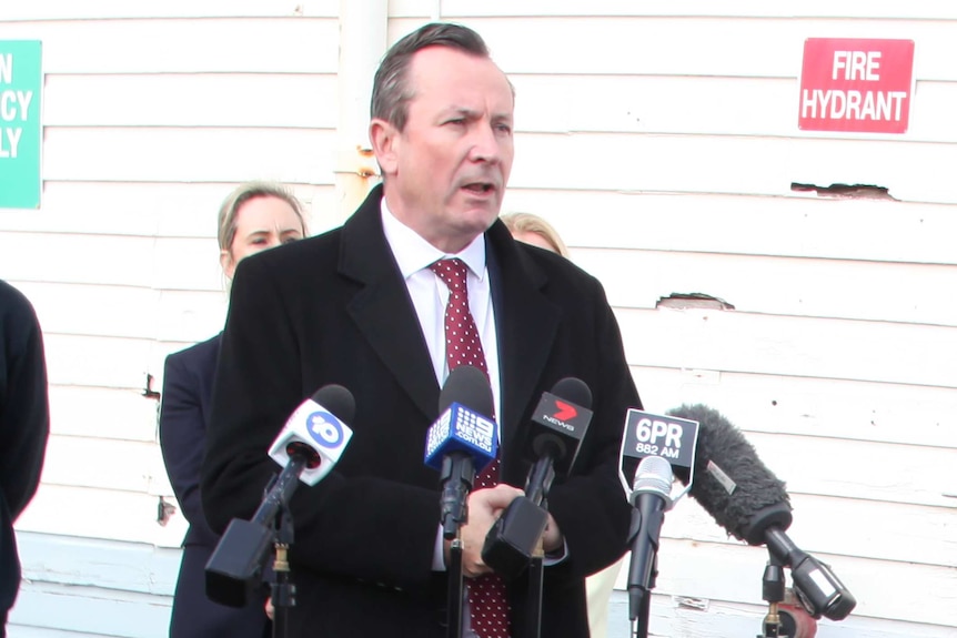 A mid shot of WA Premier Mark McGowan speaking into microphones at a media conference wearing a black coat and red tie.