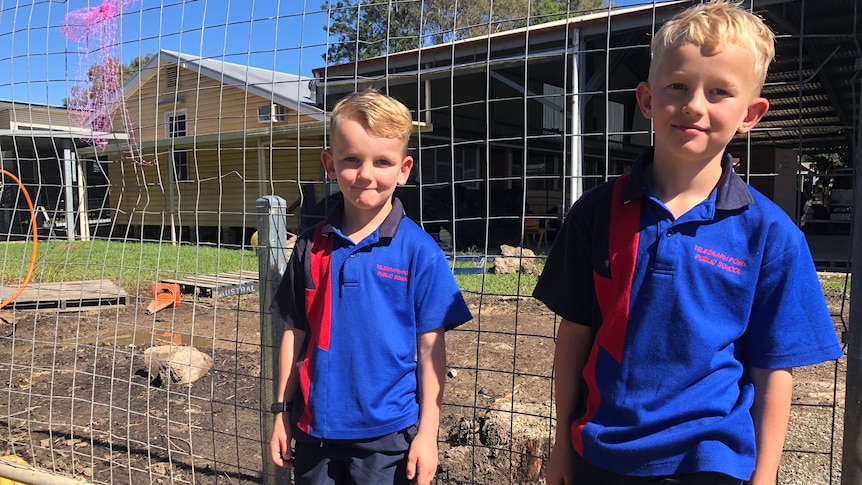 Two young blond boys in school uniform stand in front of cyclone fencing near a construction site.