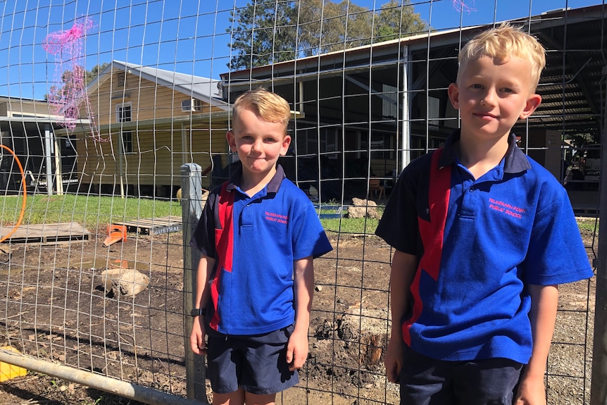 Two young blond boys in school uniform stand in front of cyclone fencing near a construction site.