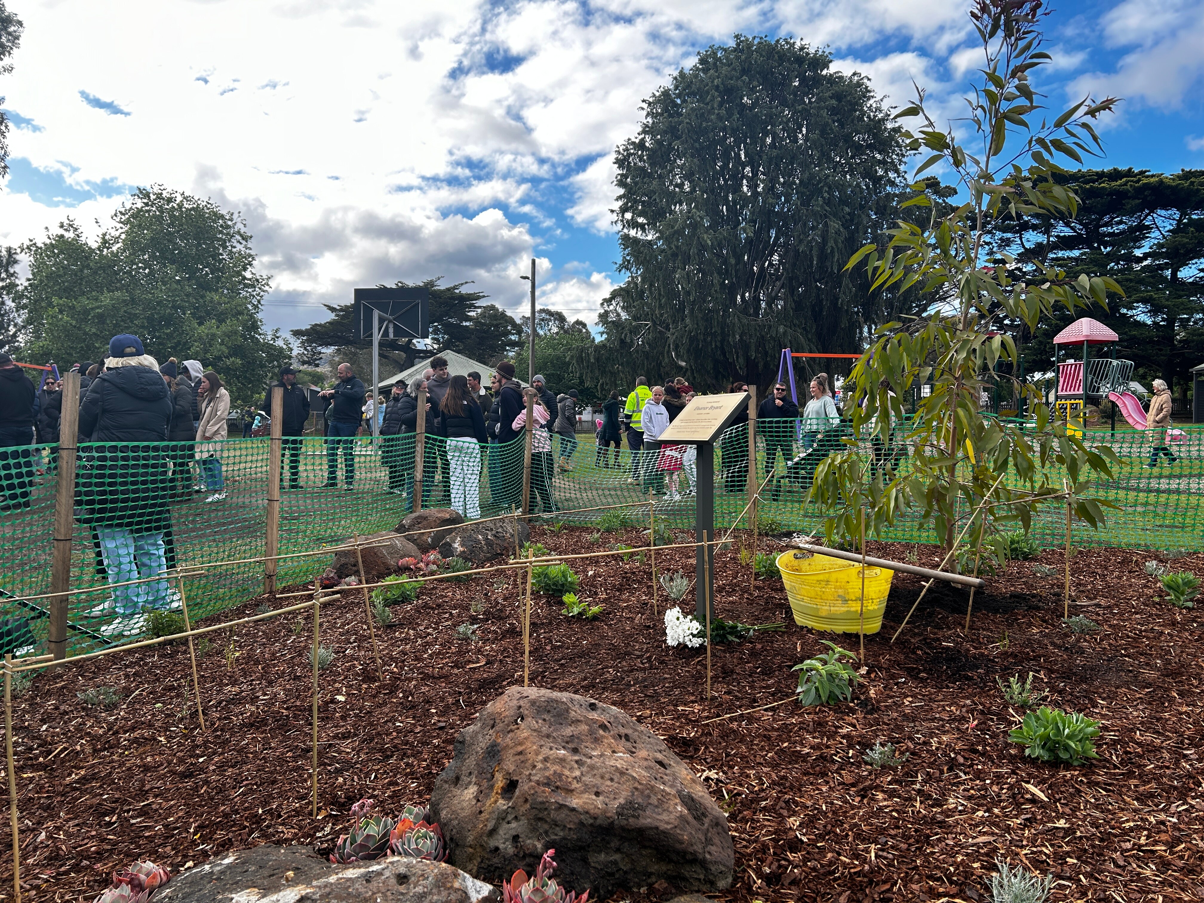Uma placa e uma pequena árvore rodeada de fita e diversas plantas. Algumas pessoas também se aglomeram em torno dele. Nuvens no céu.