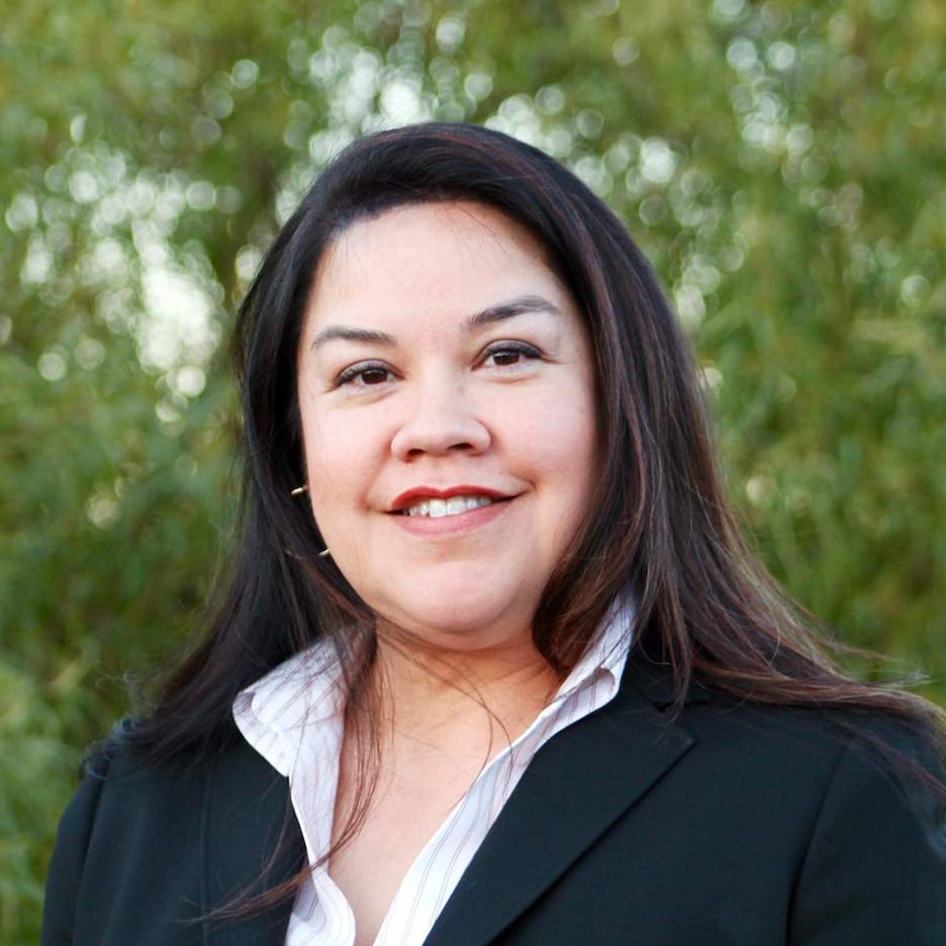 You view a close up of Christina Andrews in a black  blazer and open white shirt, standing in front of green foliage.