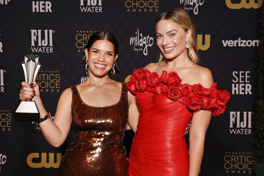 America Ferrera in a born evening gown clutching an award and Margot Robbie in a red evening gown, both smiling