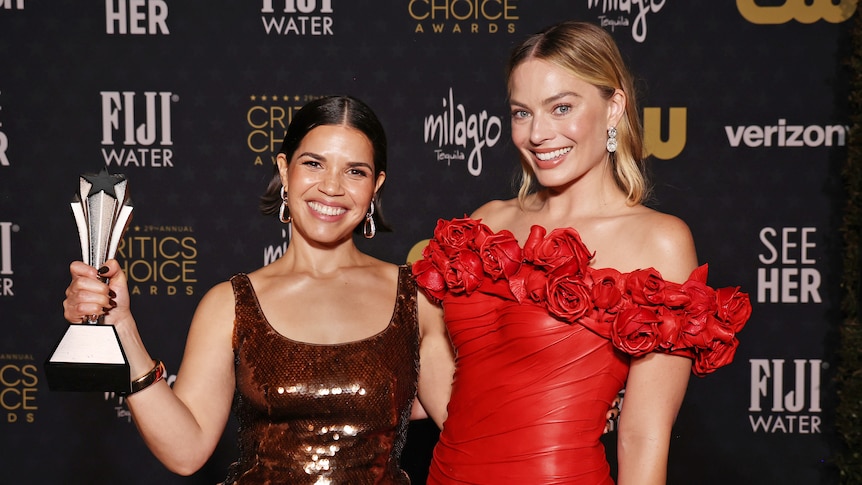 America Ferrera in a born evening gown clutching an award and Margot Robbie in a red evening gown, both smiling