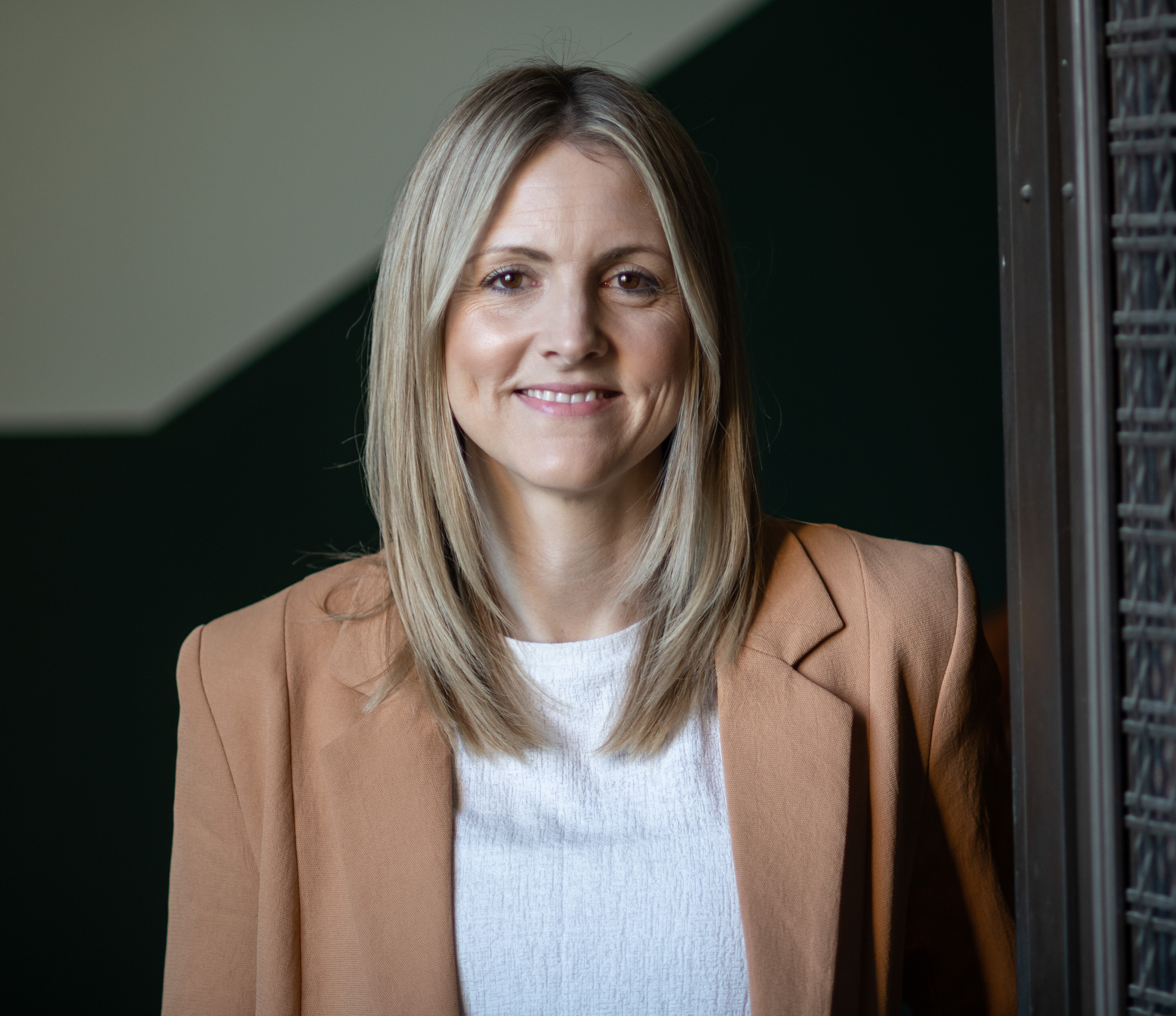 A woman in a beige jacket with long blonde hair smiles at the camera.