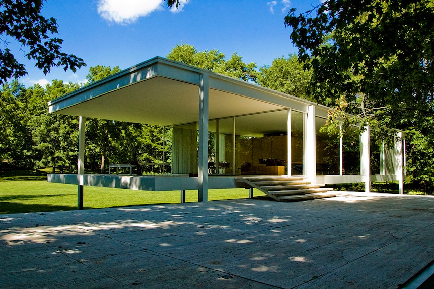 A rectangular, white modernist house is viewed on a clear day situated behind large trees that cast shadows over it.