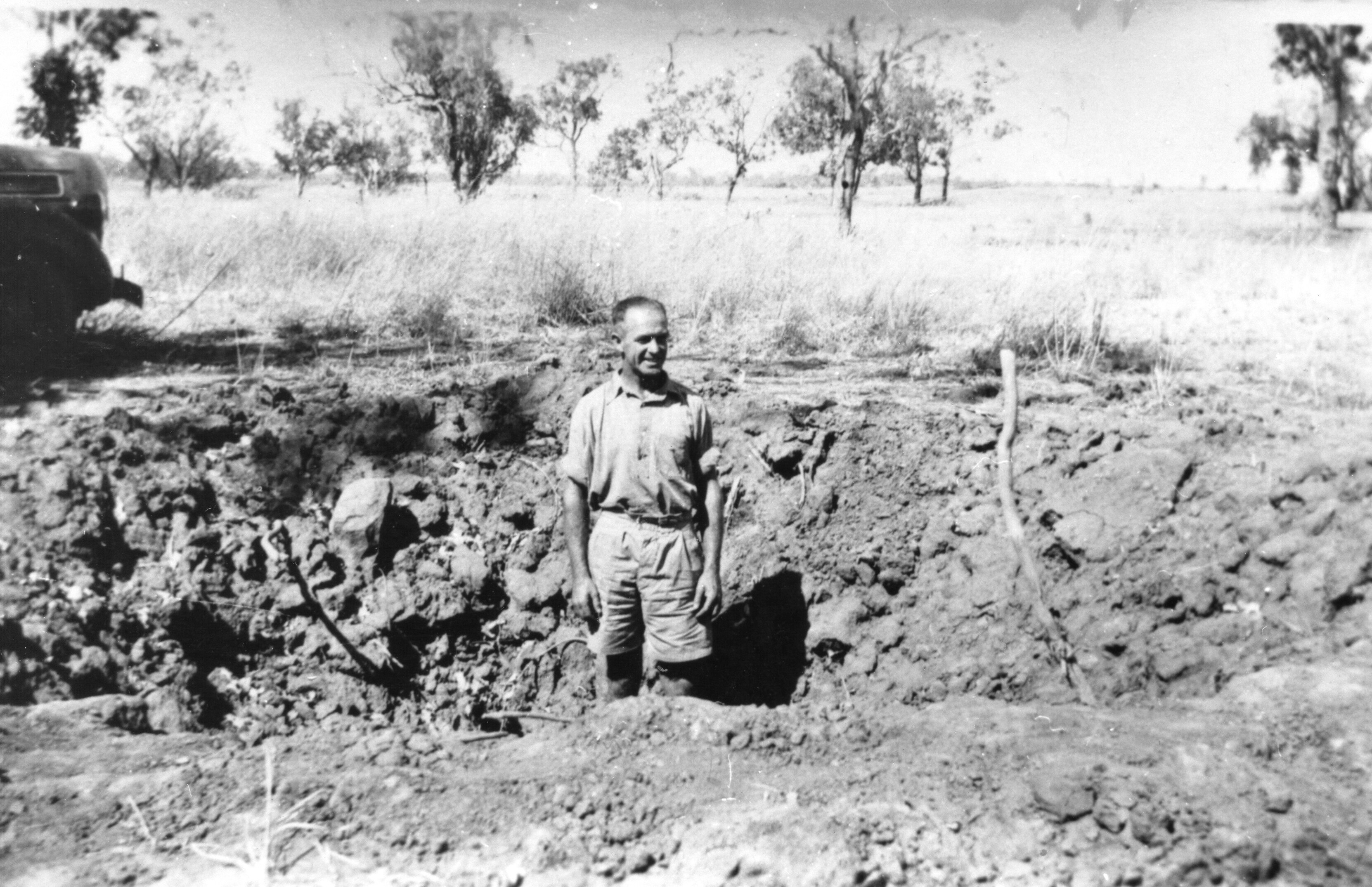 A man stands in a deep hole of rocky ground, in black and white