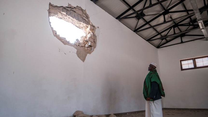 Ethiopian man looks up at large hole in building wall with bricks and rubble on the ground