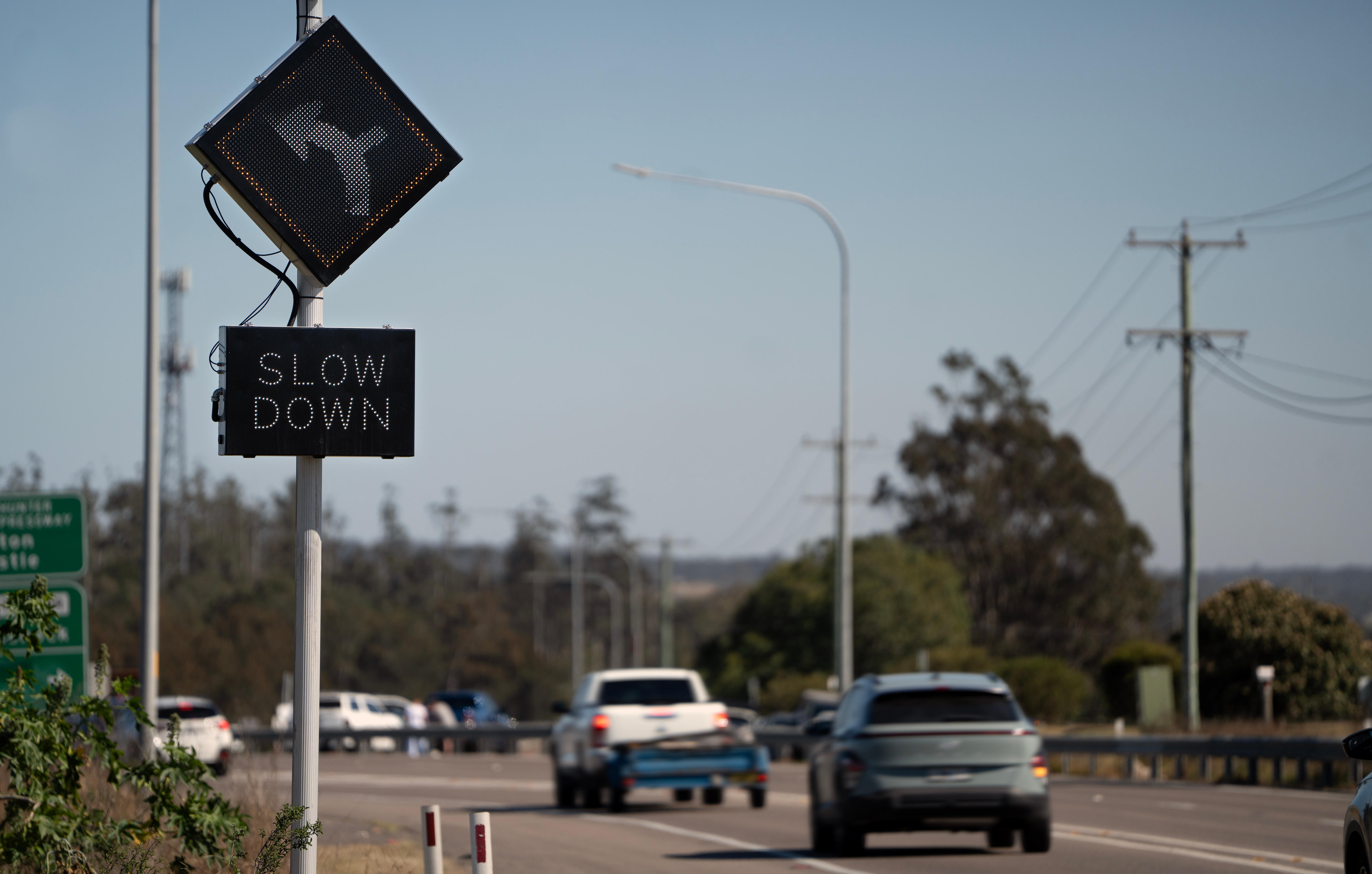 Cars travel on a road that is lined with trees. A traffic sign in foreground flashes 'Slow down'.
