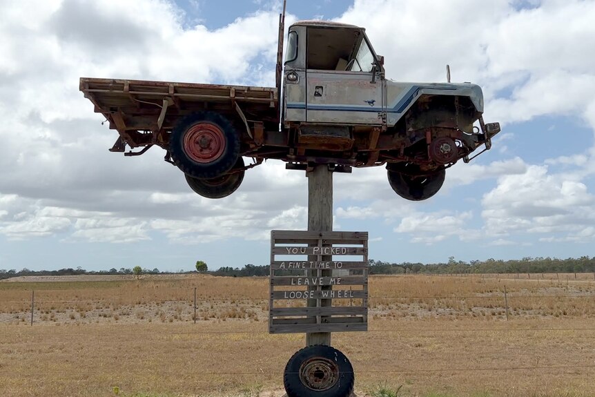 An old ute on a shortened power pole, with a sign written on a pallet and a wheel underneath.