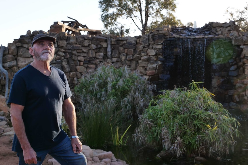 A man in a dark blue shirt and jeans stands in front of a large waterfall with reeds in its pond.