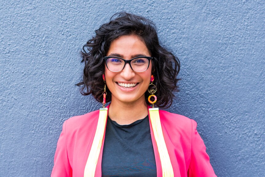 Mariam Mohammed stands in front of a blue wall. She's smiling, wearing glasses, a bright pink jacket, black tee and yellow pants