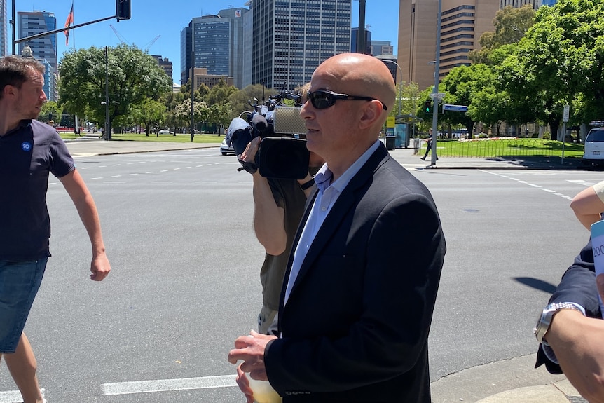 A man with a bald head wearing a suit and sunglasses walks down a street with media crews around him