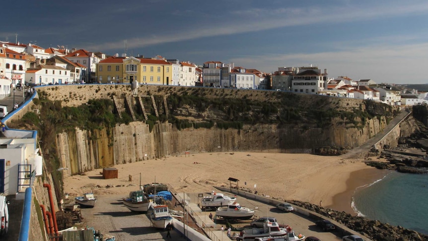 A wide photo shows a cliff face with houses behind it and a beach below.