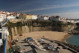 A wide photo shows a cliff face with houses behind it and a beach below.