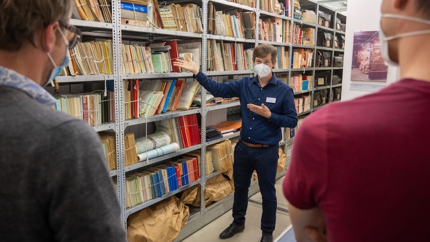 A man showing two others around a large bookshelf holding many colourful files.