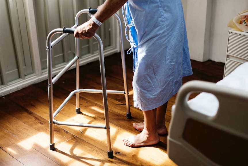A man in a hospital gown stands up with a walking frame