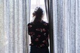 A woman stands looking out a bright window, next to her on either side are patterned floor to ceiling curtains.