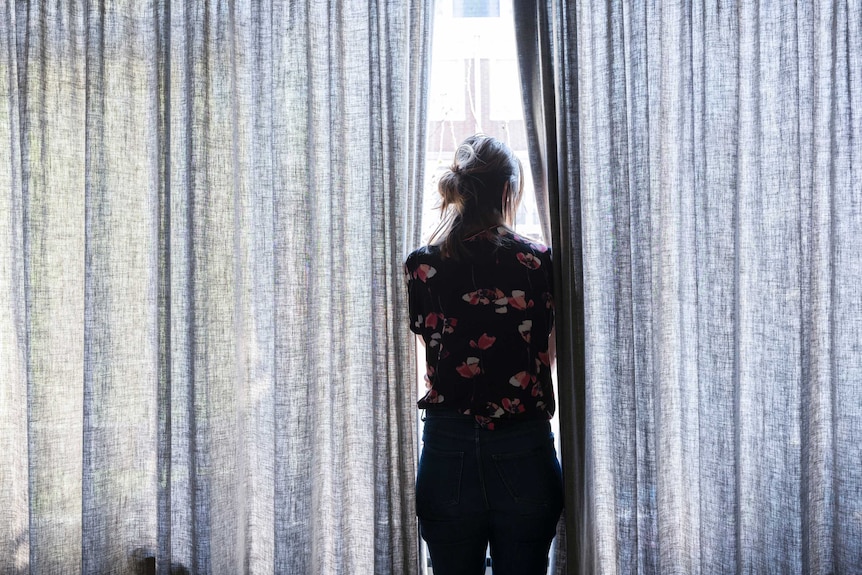 A woman stands looking out a bright window, next to her on either side are patterned floor to ceiling curtains.