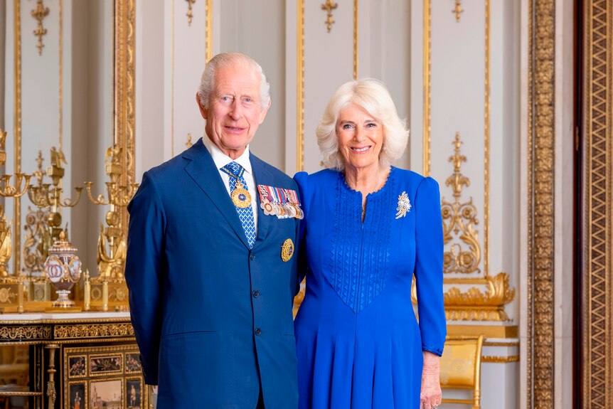 King Charles and Queen Camilla standing for a photo. Charles is wearing a blue suit, Camilla is wearing a long blue dress