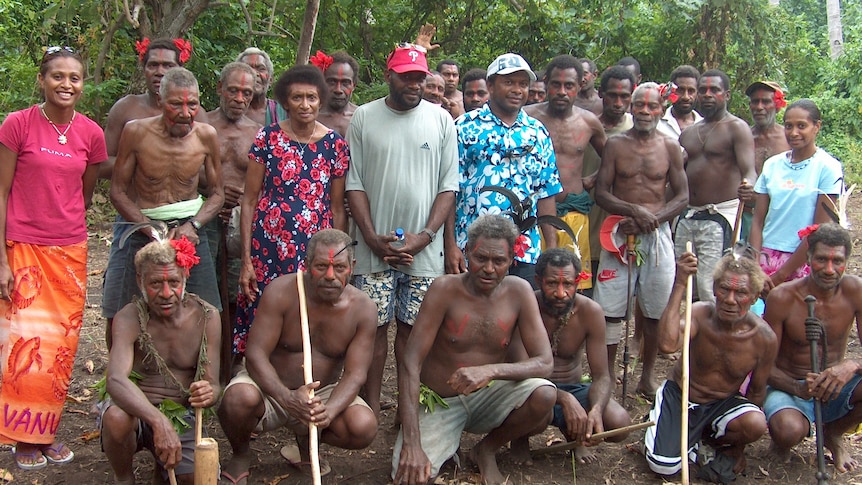 A group of people stand together looking at the camera. Some of the men are shirtless.