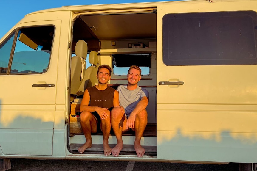 Two men sit side by side in their van smiling.