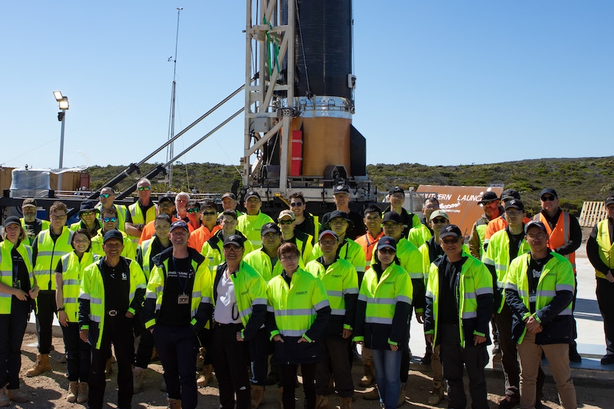 A group of people in green high vis vests stand in front of a rocket