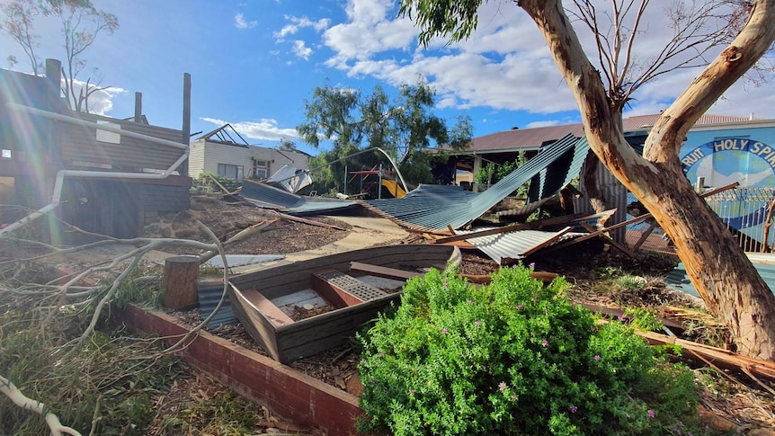 Photo of cyclone damaged school showing sheets of metal and trees uprooted with a partly destroyed house in the back.
