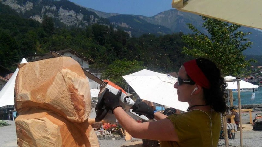 A woman carving a block of wood with a chainsaw in front of mountains
