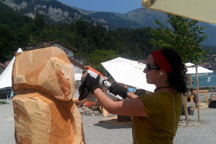 A woman carving a block of wood with a chainsaw in front of mountains
