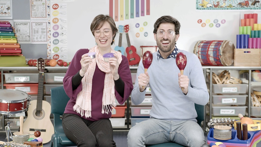 Two smiling primary school teachers sit in a decorated music classroom playing maracas and shakers