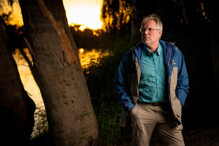 A man stands with hands in his pocket with a serious expression with a river behind him at sunset